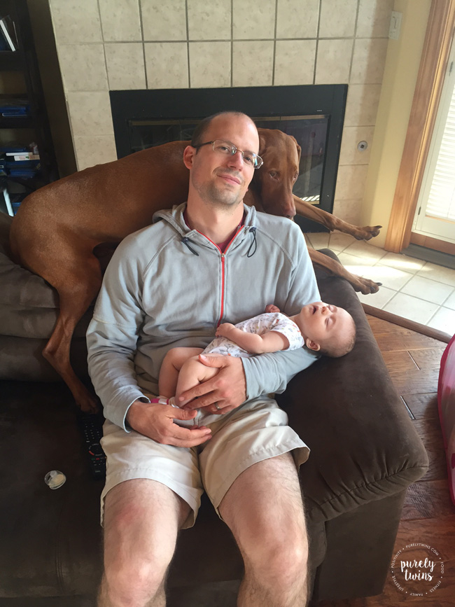 Father holding baby girl while taking a nap with Vizsla joining them on sofa. 